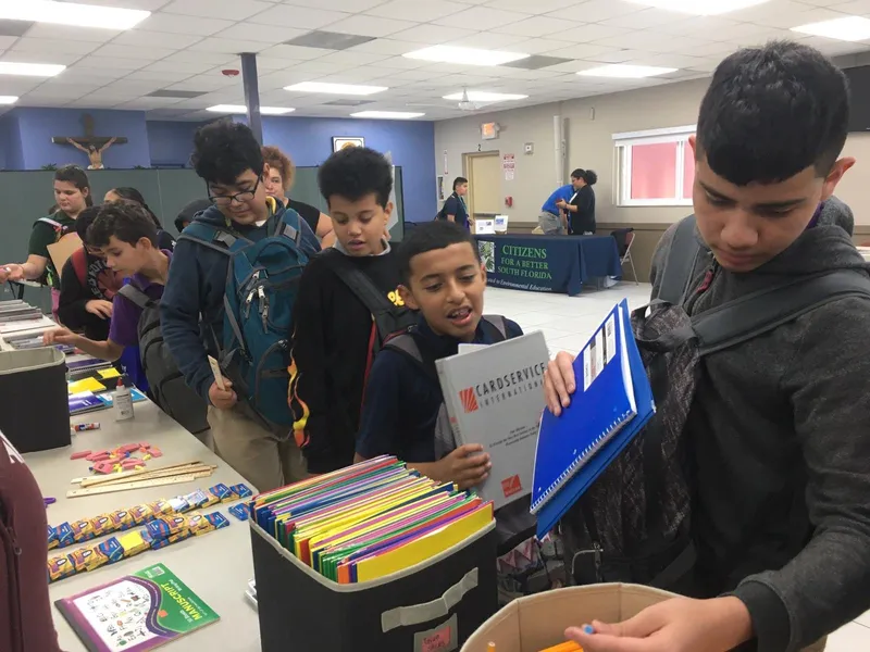 Students choose folders and supplies from boxes at a bookbag distribution table.