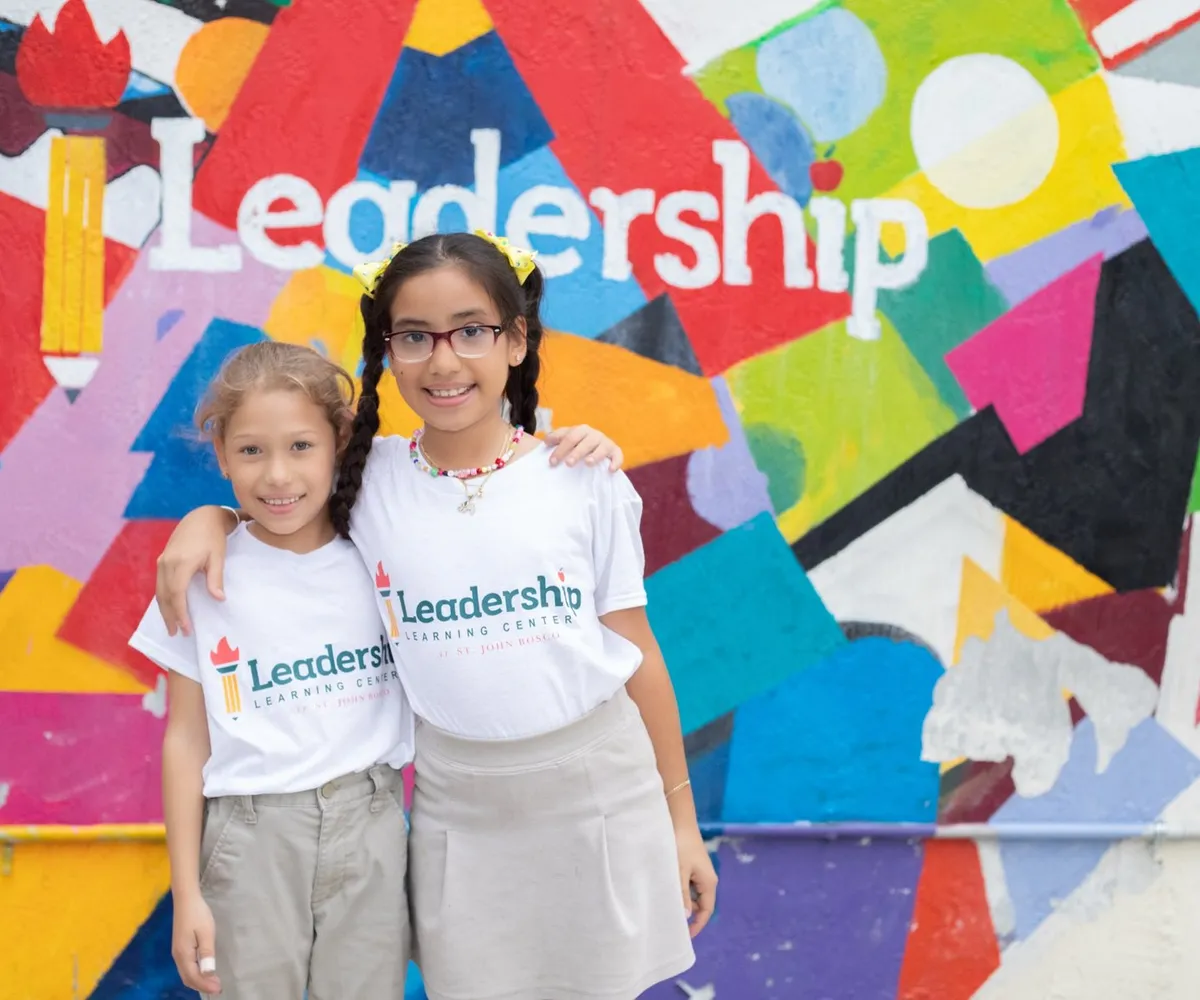 Two smiling students in Leadership Learning Center shirts stand arm in arm in front of the center’s colorful mural.