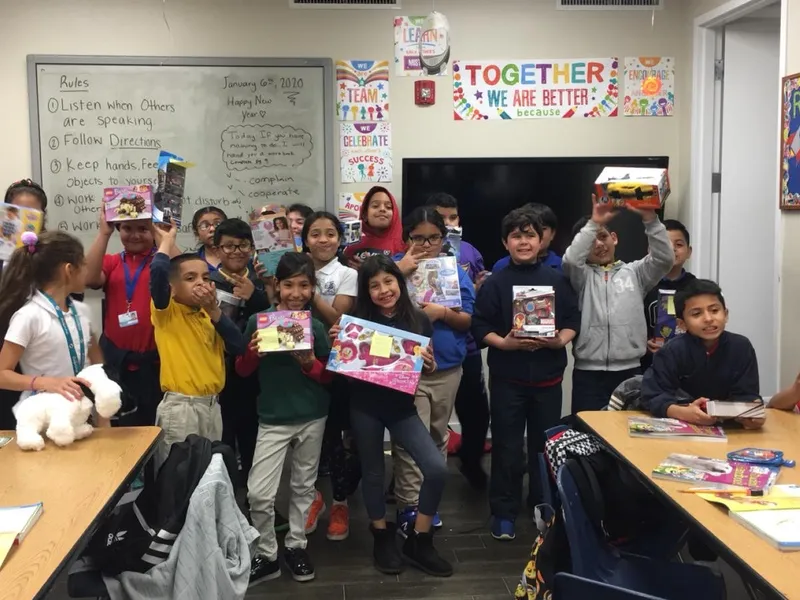 A classroom of students hold up wrapped gifts and toys at a holiday party.