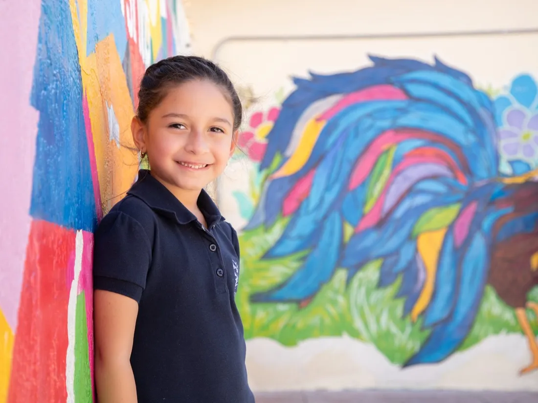 A young student smiles beside a painted rooster on the center’s mural.