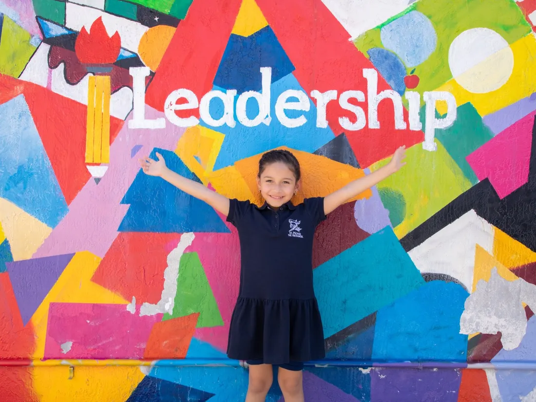 A student in a Leadership Learning Center shirt smiles with arms outstretched in front of the colorful mural.
