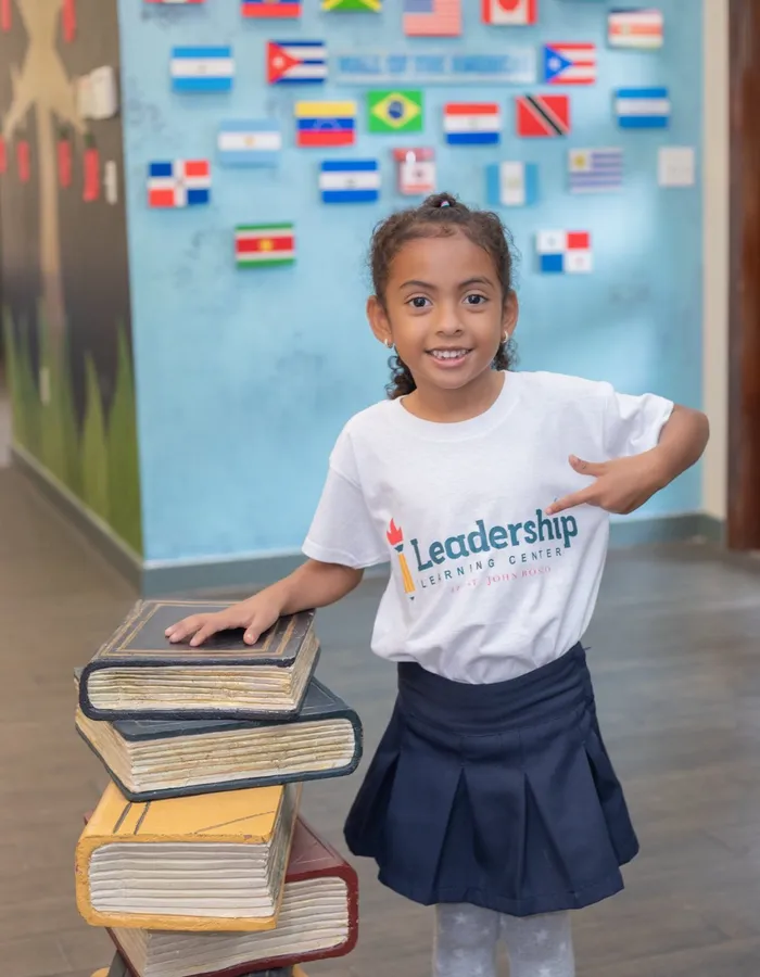 A smiling student in a Leadership Learning Center shirt rests a hand on a stack of books in front of the Wall of the Americas.